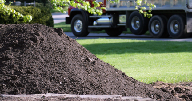 Pile of dark landscaping soil delivered on a lawn, with a dump truck parked in the background ready for a landscaping project.